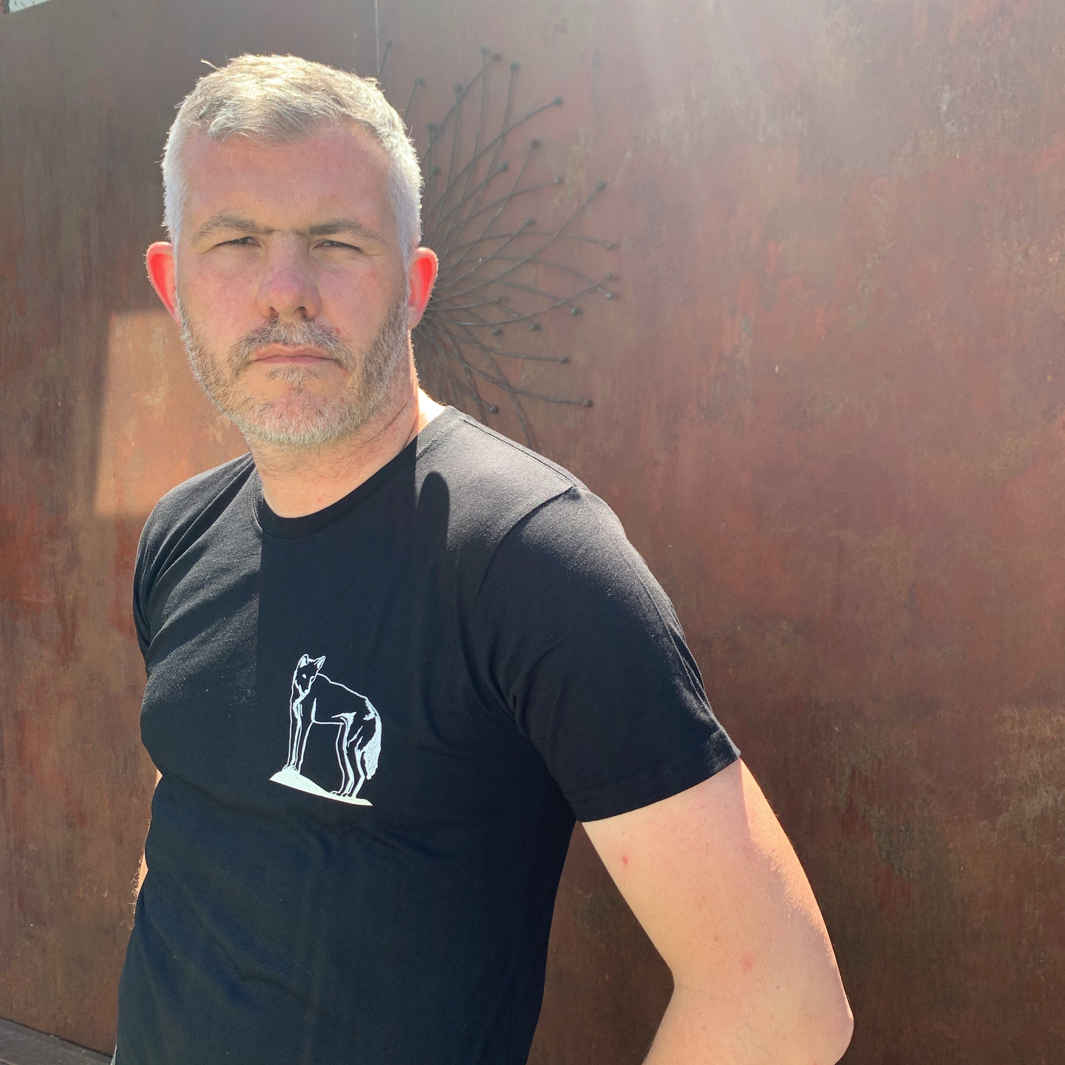 Man wearing a black t-shirt with a white dingo design against a textured wall.